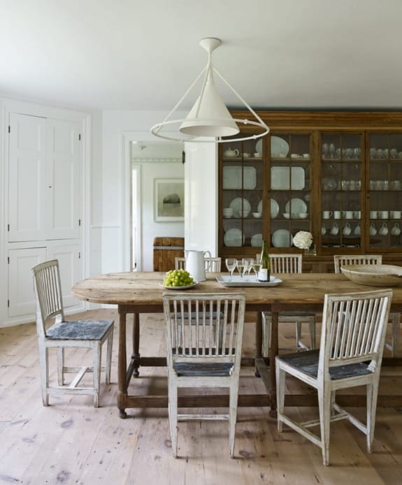 Rustic dining room with a wooden table and vintage chairs, surrounded by white walls and a large cabinet filled with plates.