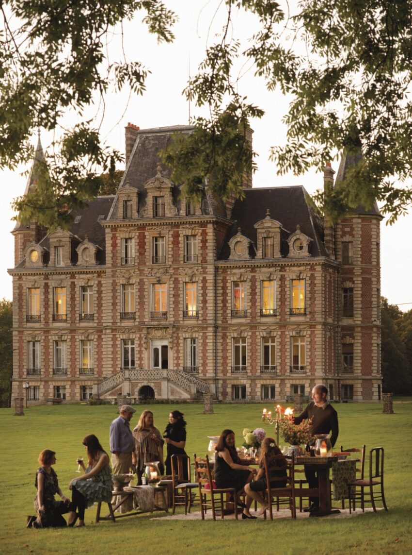 Group of people dining outdoors with a large castle-like building in the background surrounded by greenery.