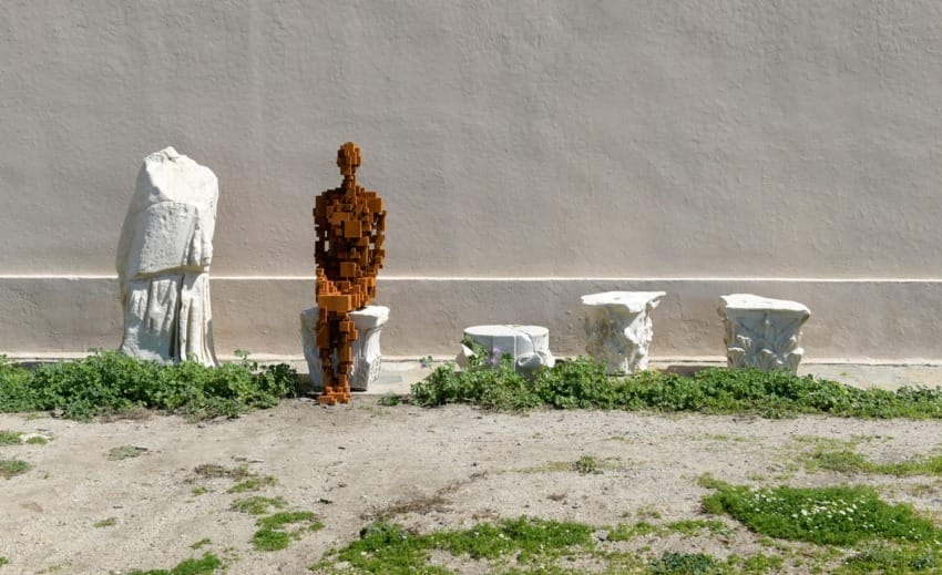 Modern sculpture of a faceless human among broken classical statues against a beige wall and green grass.