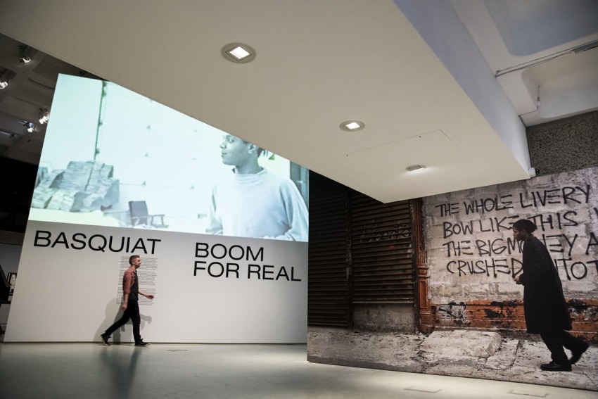 Exhibition with large images and text "Basquiat Boom For Real" on the walls, person walking by.