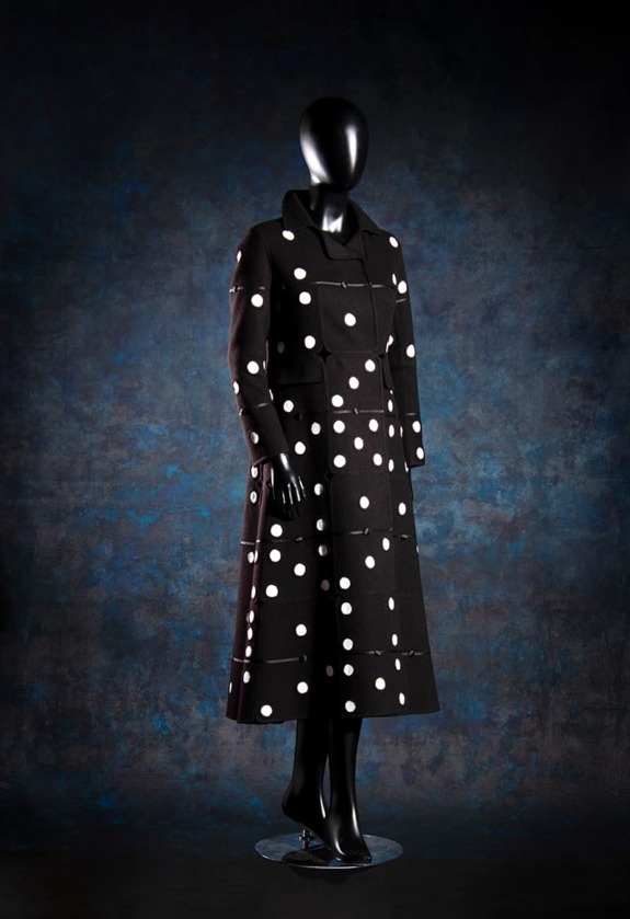 Mannequin wearing a long black coat with white polka dots against a dark blue and black background.