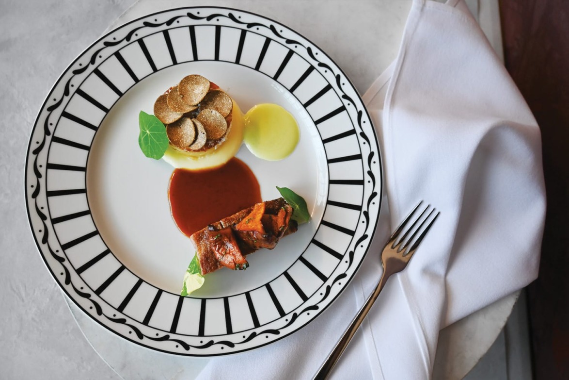 Gourmet dish with steak, sauce, potato cake topped with truffle slices on an elegant black and white plate near a fork.
