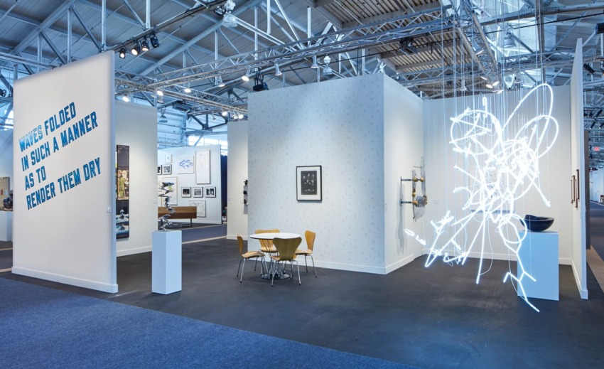 Art exhibition with neon light sculpture, contemporary artworks, and seating area inside a spacious industrial-style gallery.