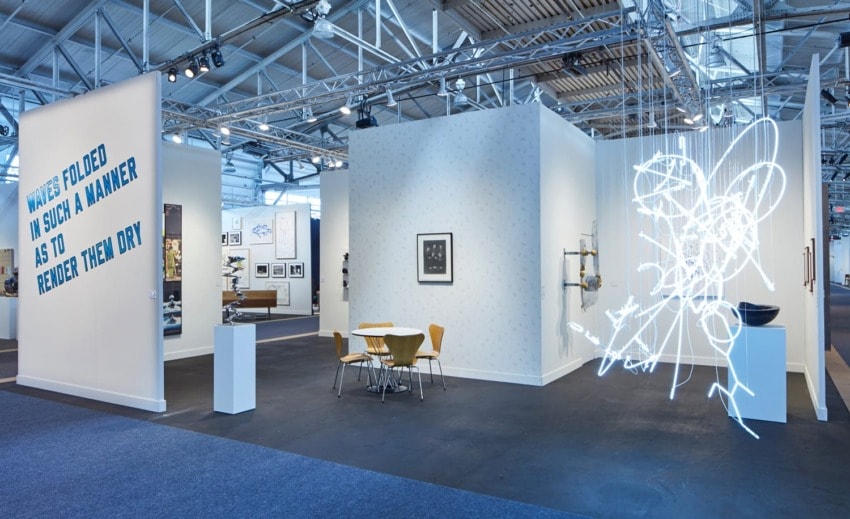 Art exhibition with neon light sculpture, contemporary artworks, and seating area inside a spacious industrial-style gallery.