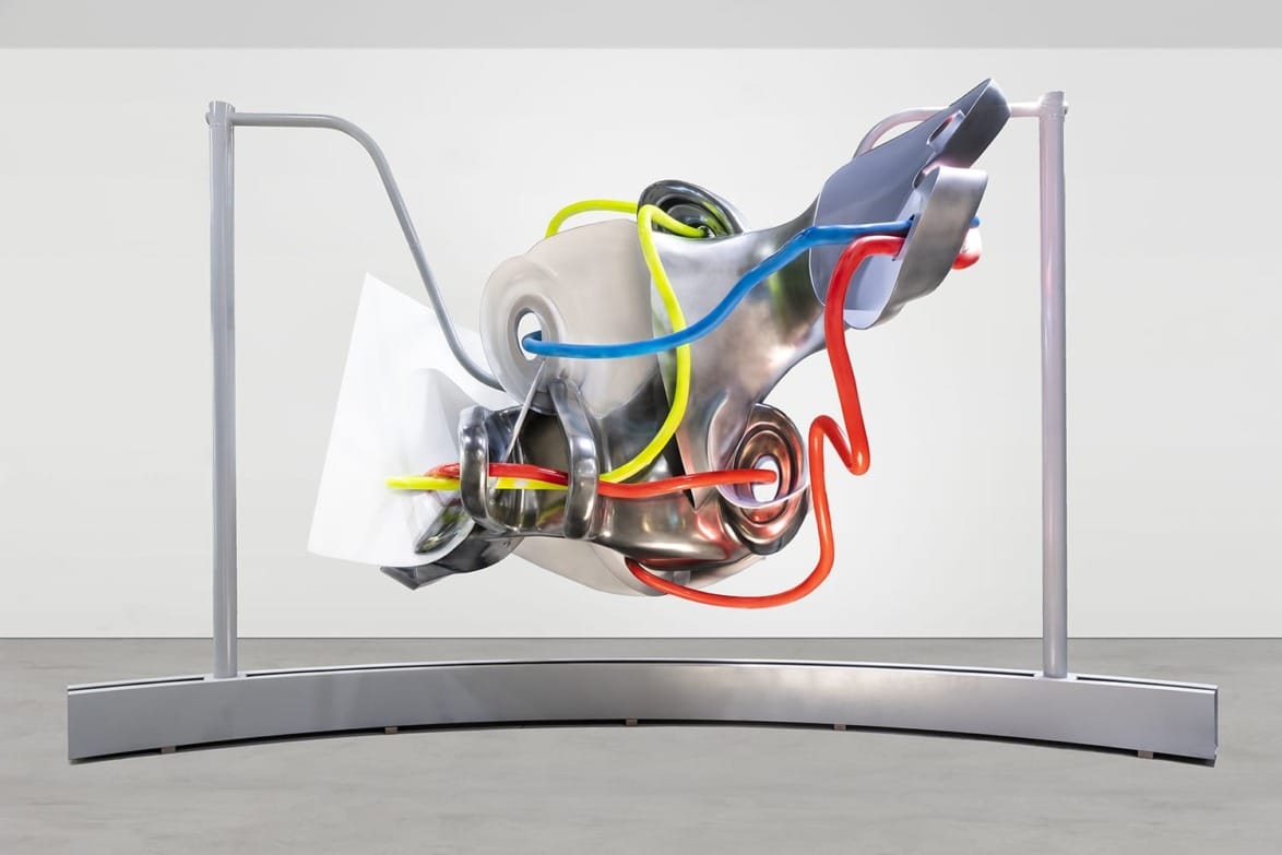 Abstract metal sculpture with colorful tubing and geometric shapes displayed on a curved base in a gallery setting.