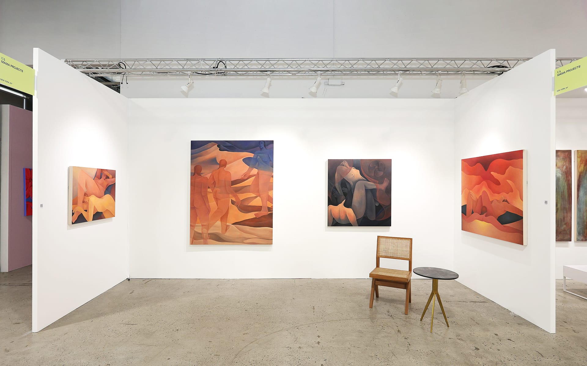 Art gallery exhibit with abstract paintings on white walls, a wooden chair, and a small round table on a concrete floor.