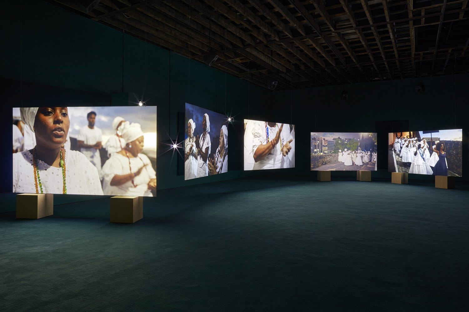 Art exhibition with multiple illuminated screens displaying images of people in white clothing in a dark room.