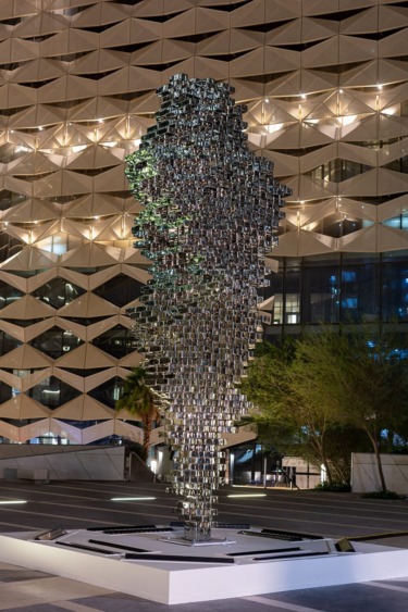 Modern abstract metal sculpture in an urban plaza at night, with a geometric building facade in the background.