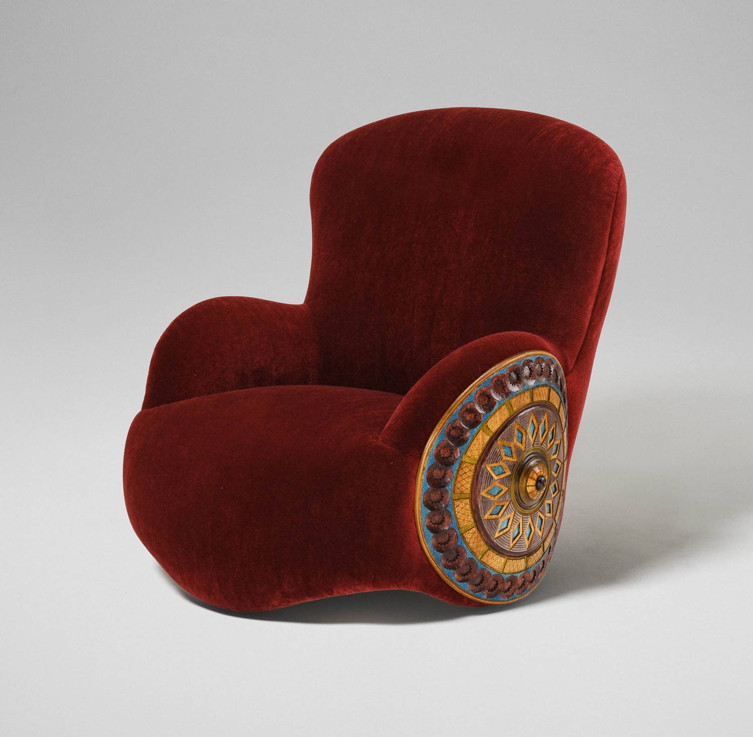 Red velvet armchair with an ornate wooden side detail featuring an intricate circular design.