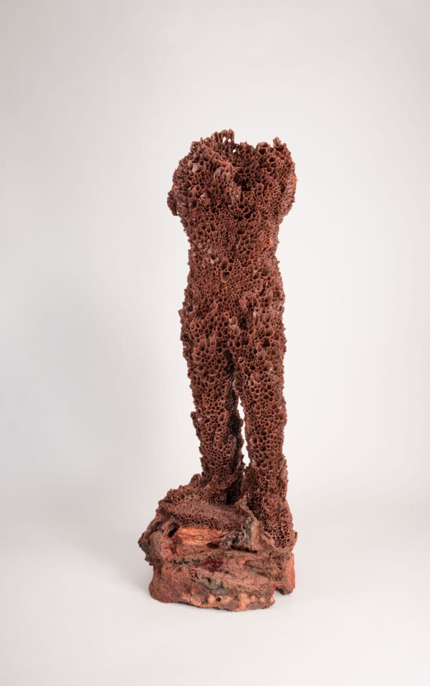 Abstract sculpture resembling a human torso made of textured, reddish material against a neutral background.