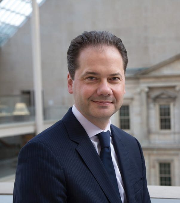 Smiling person in a dark suit and tie, standing indoors with a blurred building interior in the background.