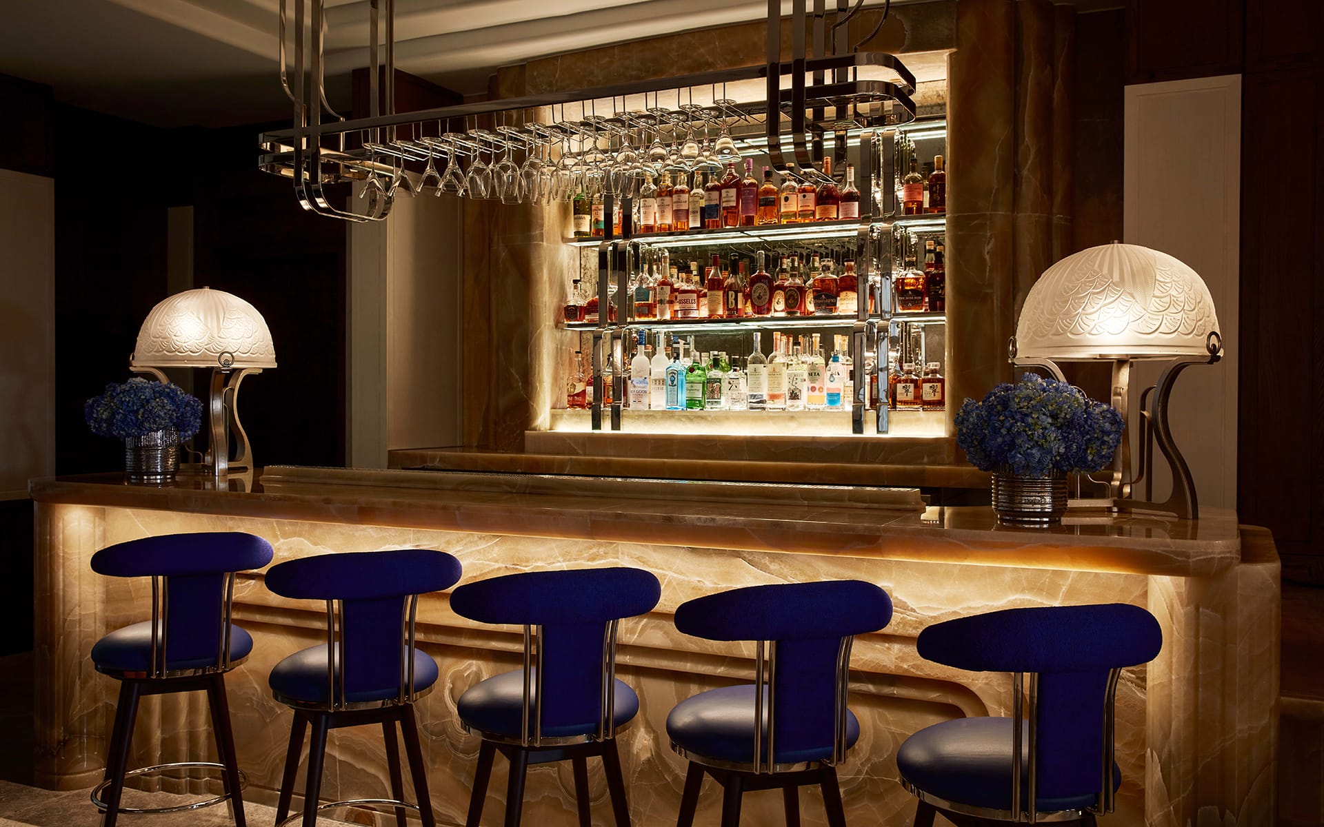 Modern bar with backlit onyx countertop, blue chairs, decorative lamps, and shelves stocked with various liquor bottles.