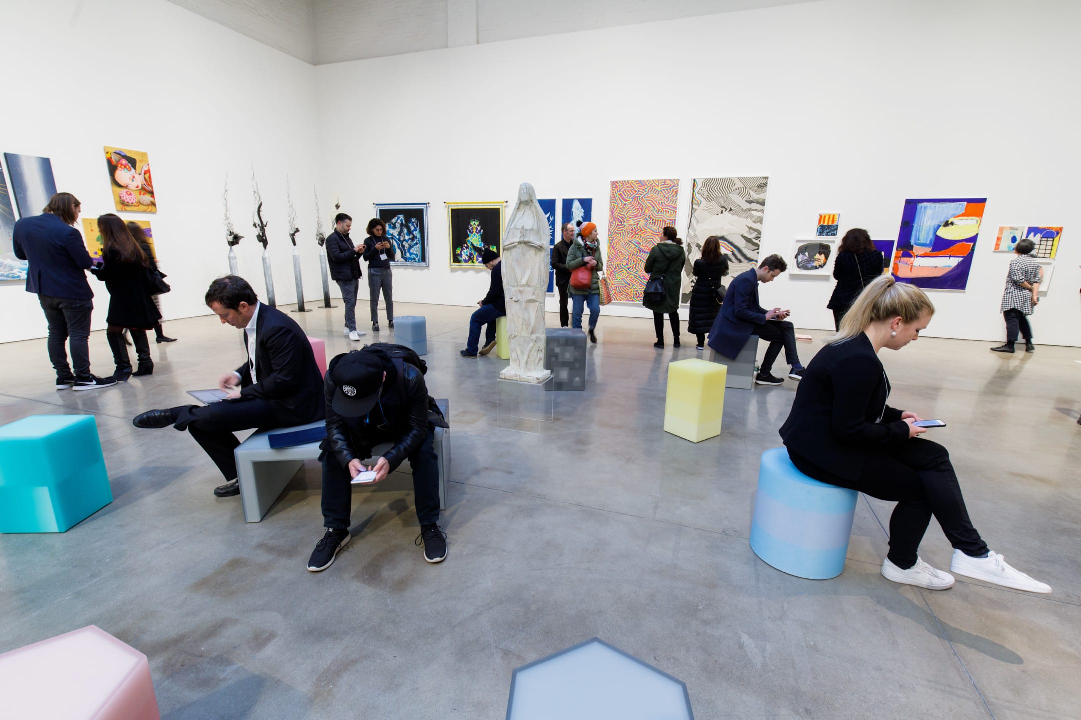 Gallery visitors observe various artworks and sculptures displayed in a spacious, well-lit art exhibition hall.