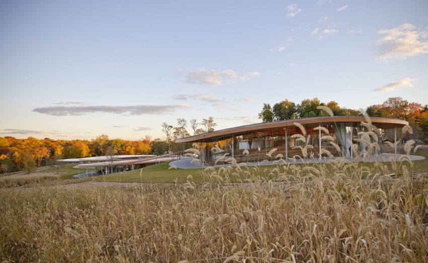 Modern building with a curved roof set in a grassy field, surrounded by colorful autumn trees under a partly cloudy sky.