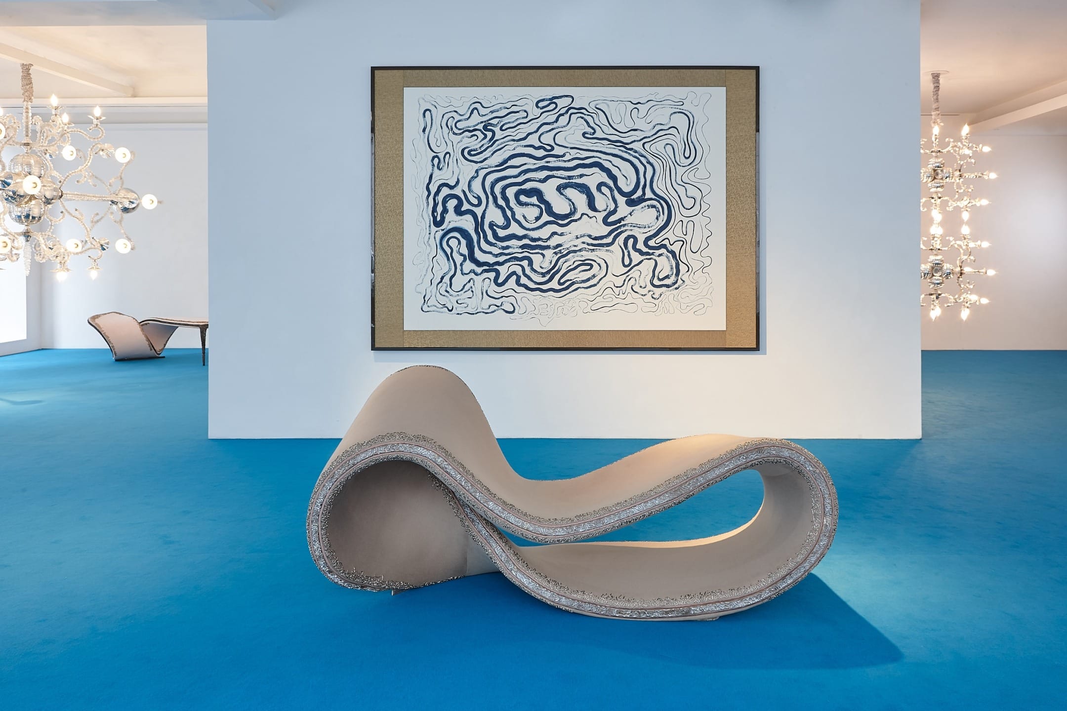 Abstract-shaped beige chair on blue floor with abstract painting on wall in a brightly lit room.