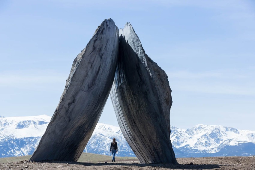Person standing beneath large split rock formation with snow-capped mountains in the background under a clear blue sky.