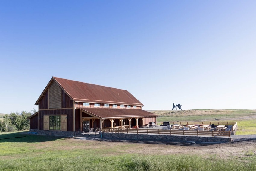 Brown barn with a sloped roof on a sunny day, surrounded by green grass and a sculpture in the distance.