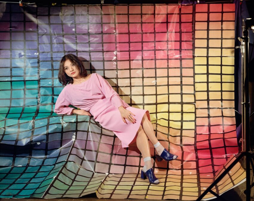 Person in a pink outfit leaning against a colorful, gradient backdrop with a grid pattern in a studio setting.