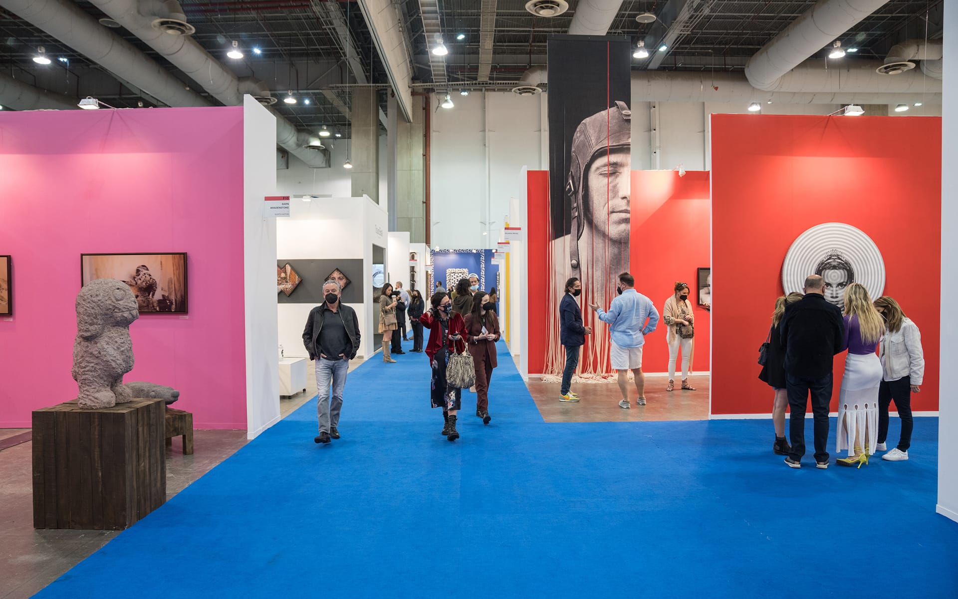 Art gallery with people exploring colorful exhibits and sculptures on blue carpeted floor.