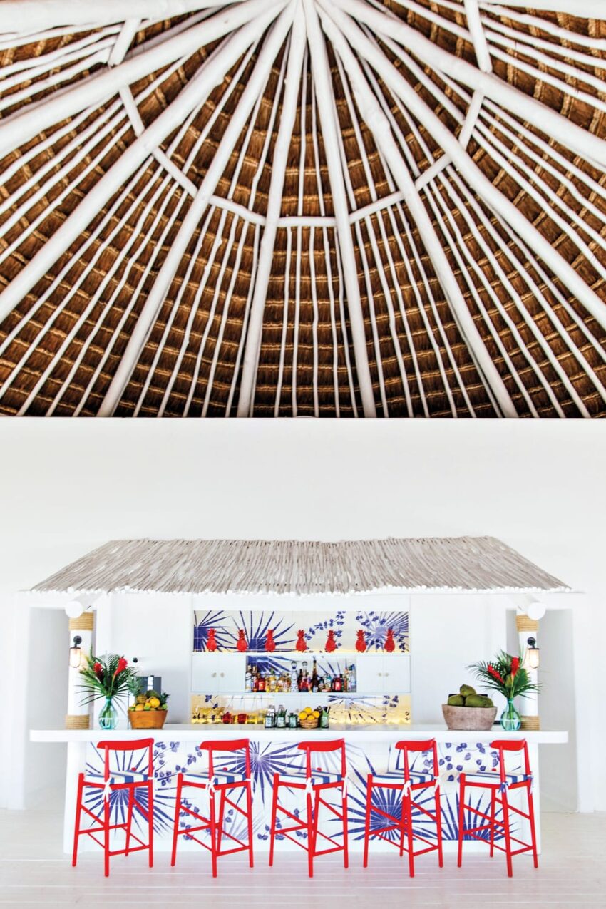 Tropical bar with a thatched roof and vibrant red chairs, decorated with colorful tiles and lush green plants.