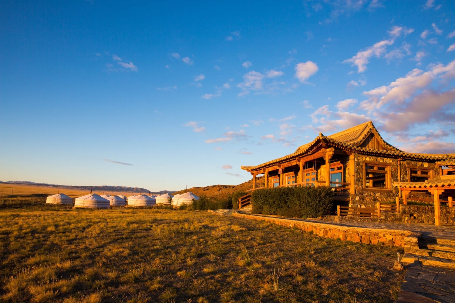 Scenic view of traditional yurts and a wooden building in a vast, sunlit grassland under a blue sky with clouds.