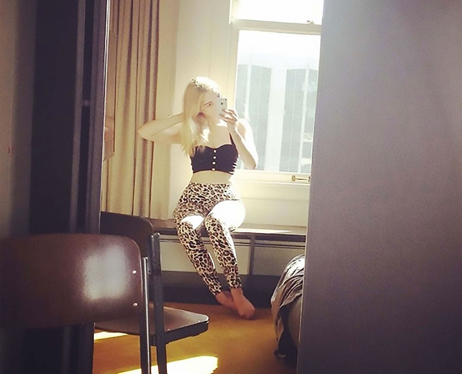 Person taking a mirror selfie in a bright room, wearing a black top and leopard print pants, with a window in the background.