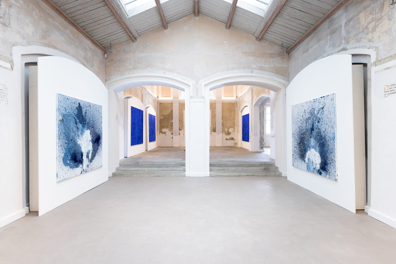 Art gallery interior with abstract blue and white paintings on white walls, natural light through skylights.