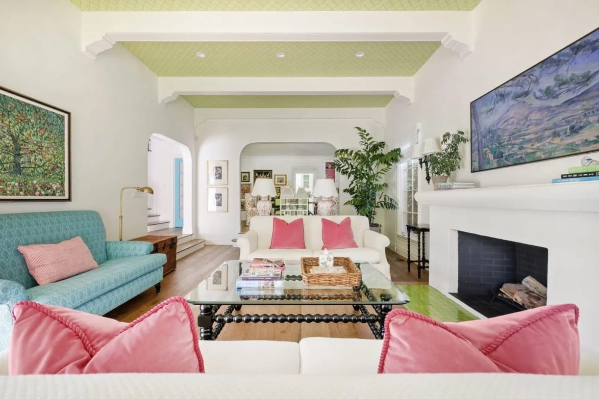 Bright living room with a white sofa, pink cushions, a green ceiling, and colorful artwork on walls above a fireplace.