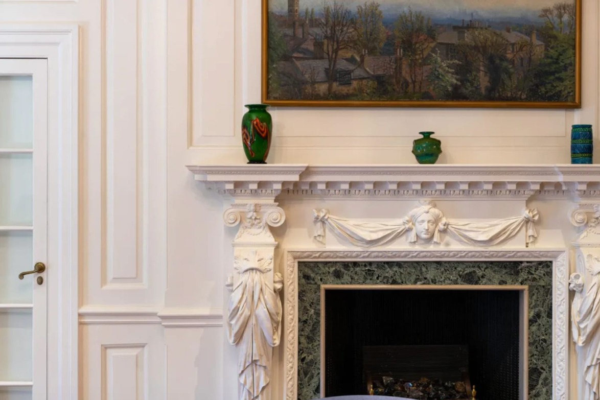 Elegant fireplace with ornate white mantel, green vases, and framed landscape painting above, set in a classic interior setting.