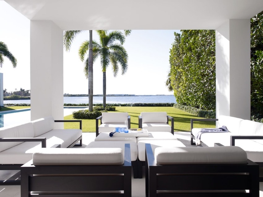Modern outdoor patio with white cushioned furniture, views of palm trees, green lawn, and distant water on a sunny day.