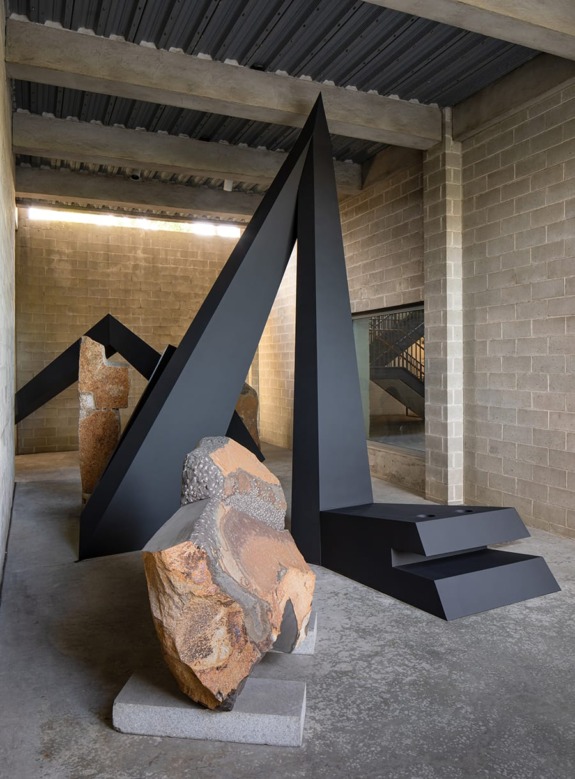 Abstract geometric sculptures with sharp angles and textured stones inside an industrial-style building interior.