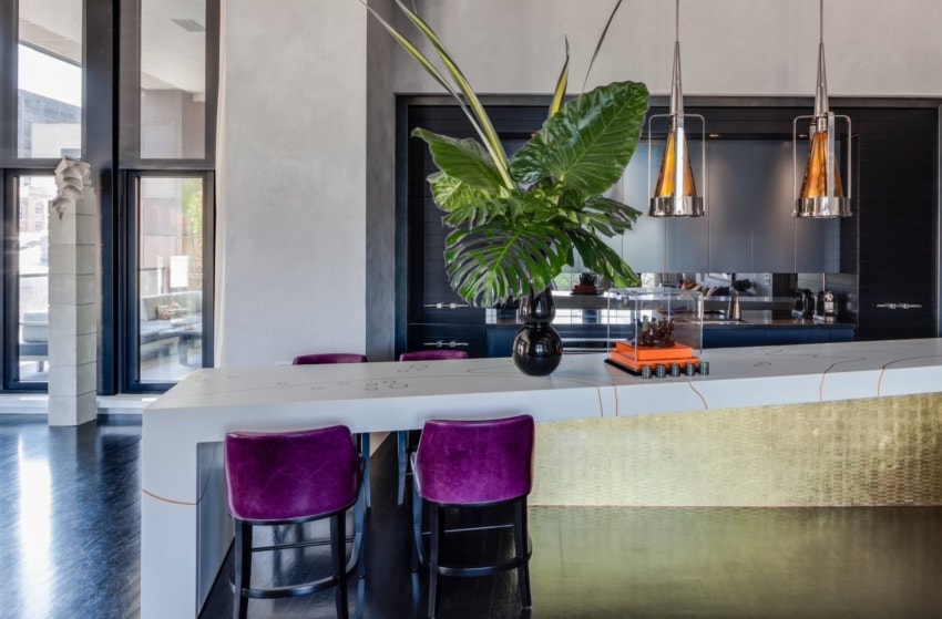 Modern kitchen with white island, purple chairs, large green plant, and pendant lights.