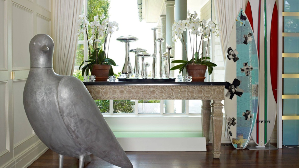 Room with large bird sculpture, ornate table with plants, shiny metal vases, and decorative surfboards by the window.