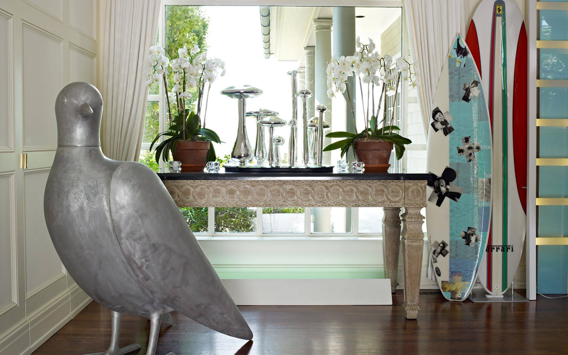 Room with large bird sculpture, ornate table with plants, shiny metal vases, and decorative surfboards by the window.