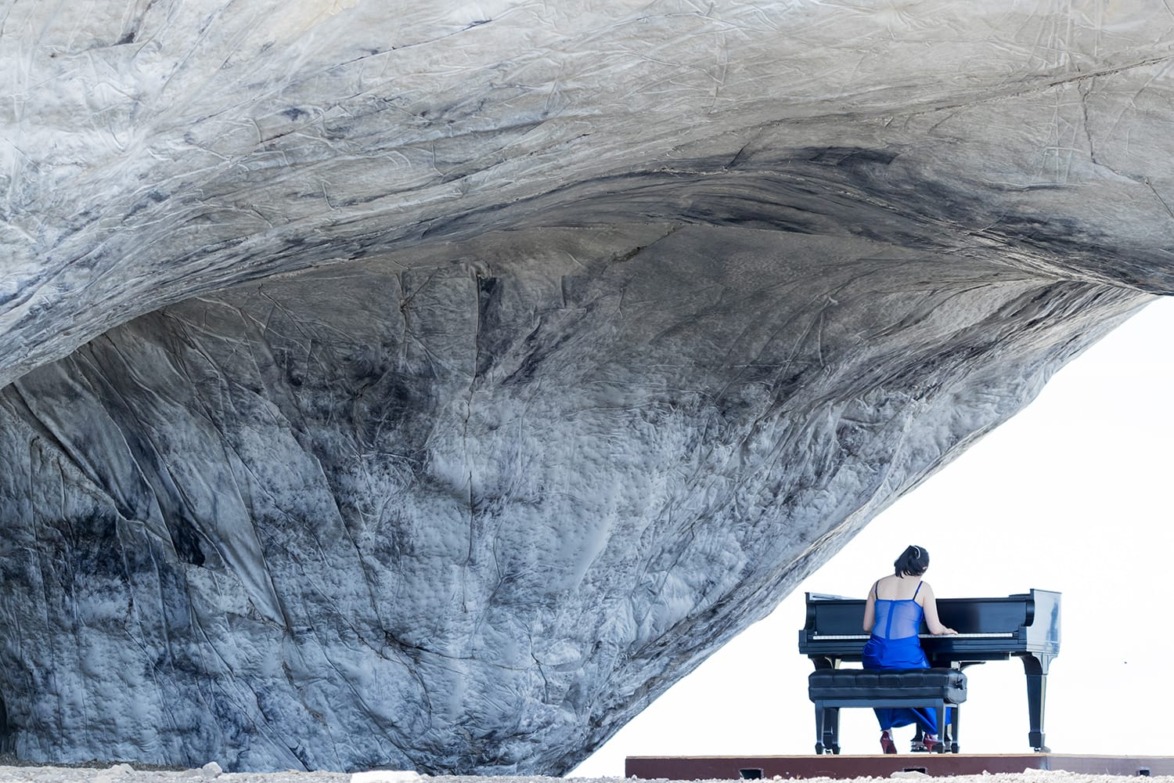 Woman in a blue dress playing a piano under a large rock formation in a vast, open landscape.