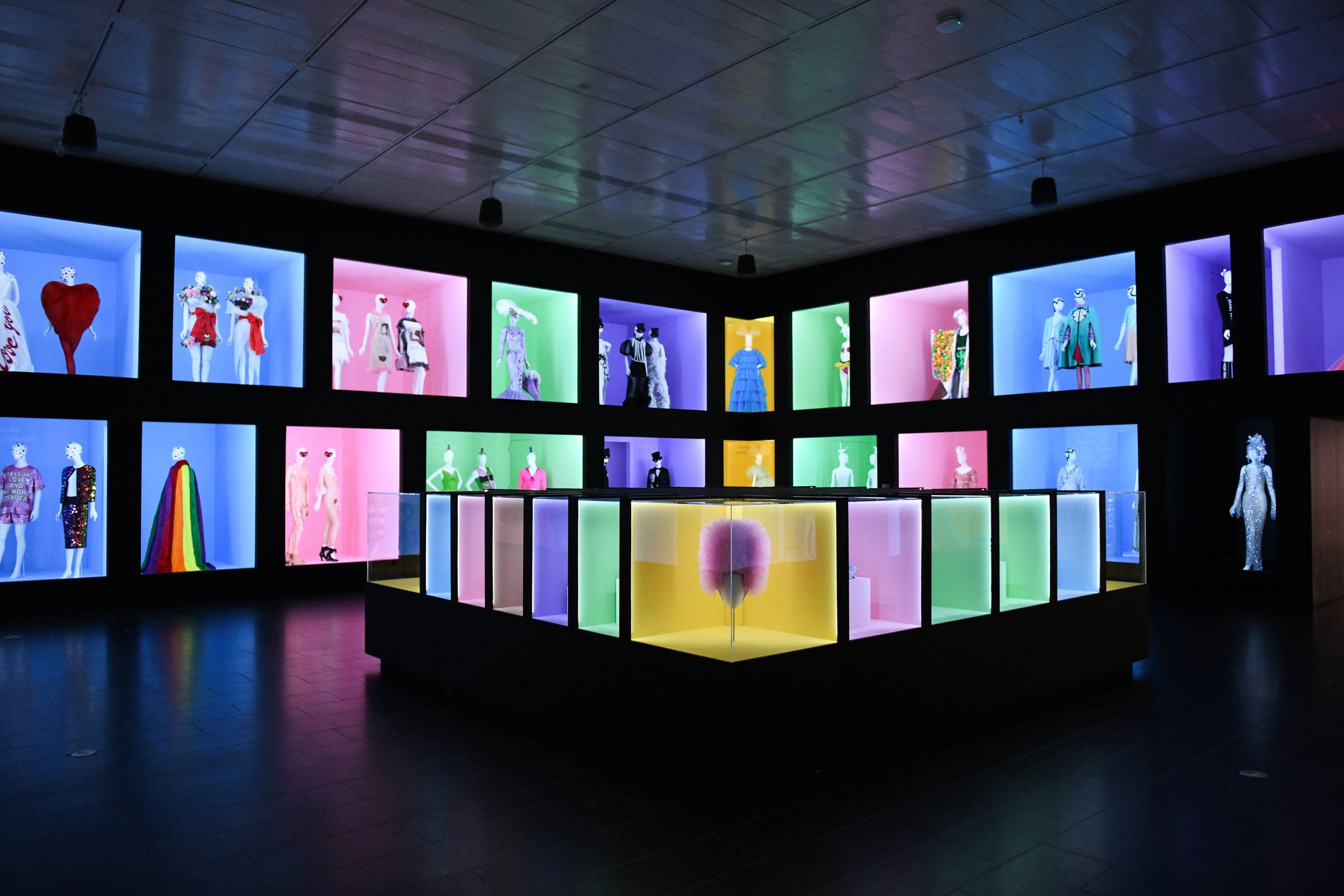 Exhibit displaying mannequins in colorful outfits set in illuminated boxes within a modern gallery space.
