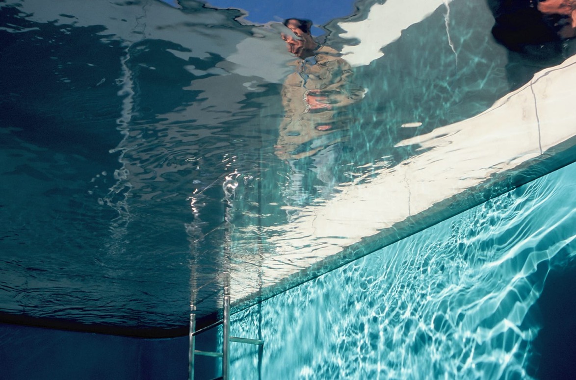 Artistic view of a swimming pool from underwater with reflections and distorted figures above, emphasizing light and water movement.