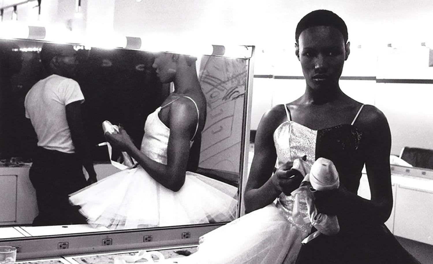 Ballet dancer in a dressing room holding pointe shoes, with costume reflected in mirror, preparing for a performance.