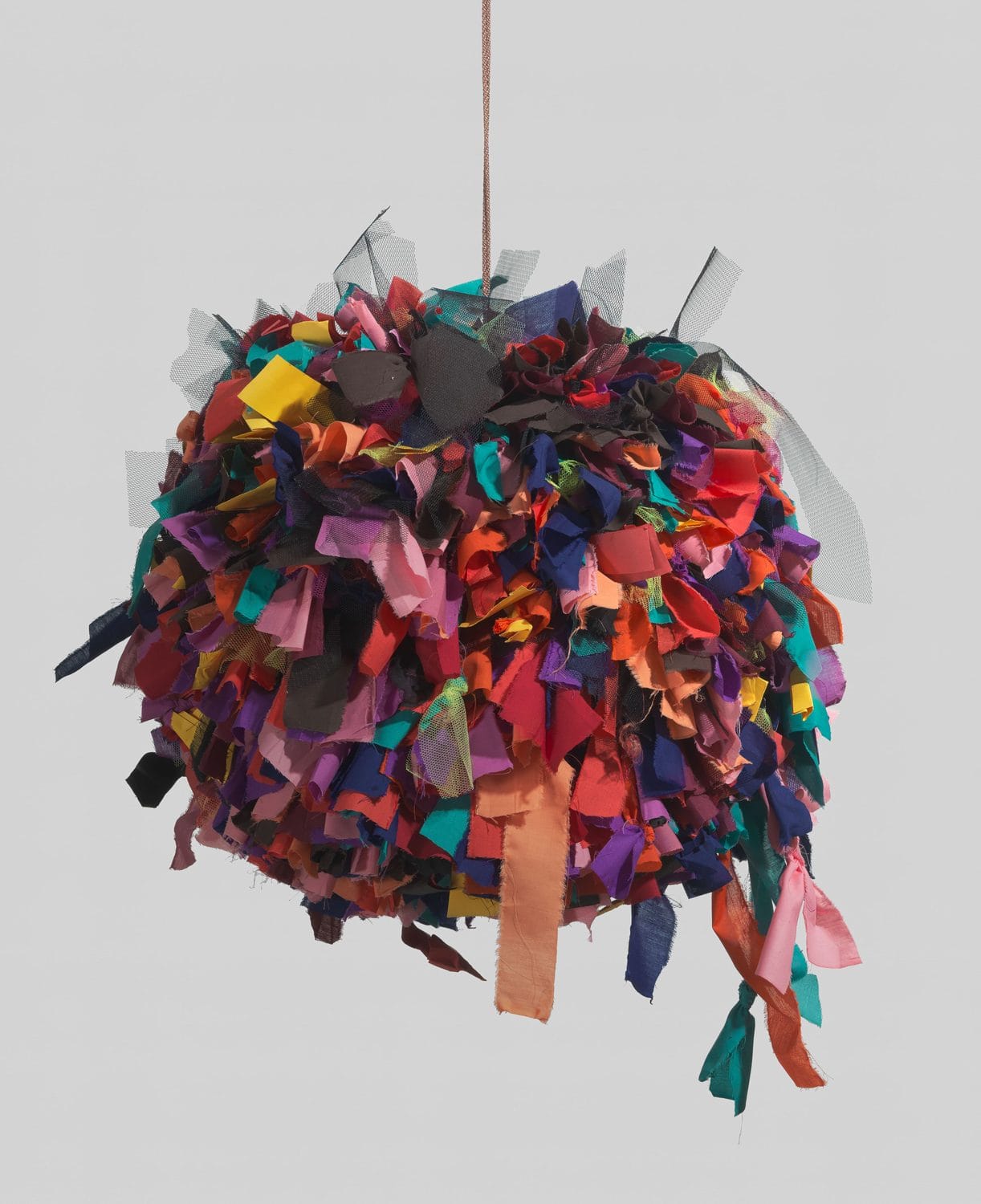 Multicolored hanging sculpture made of various fabric strips in different colors and textures against a plain background.