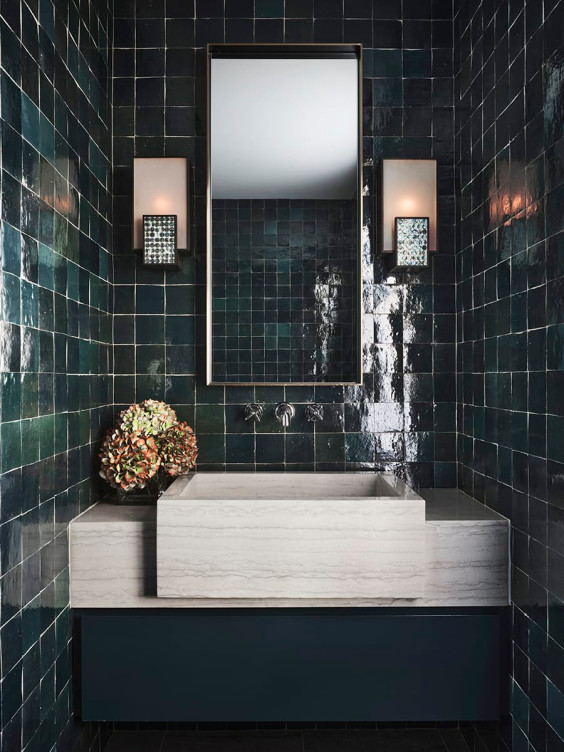 Modern bathroom with dark green tiled walls, a rectangular mirror, wall sconces, and a stone sink with decorative plant.