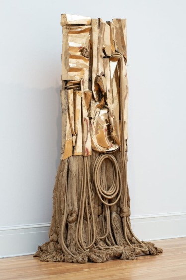 Abstract sculpture with hanging ropes and metallic strips standing against a white wall on a wooden floor.