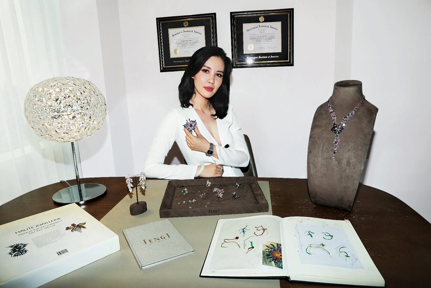 Person sitting at a desk with jewelry, awards on the wall, and open books.