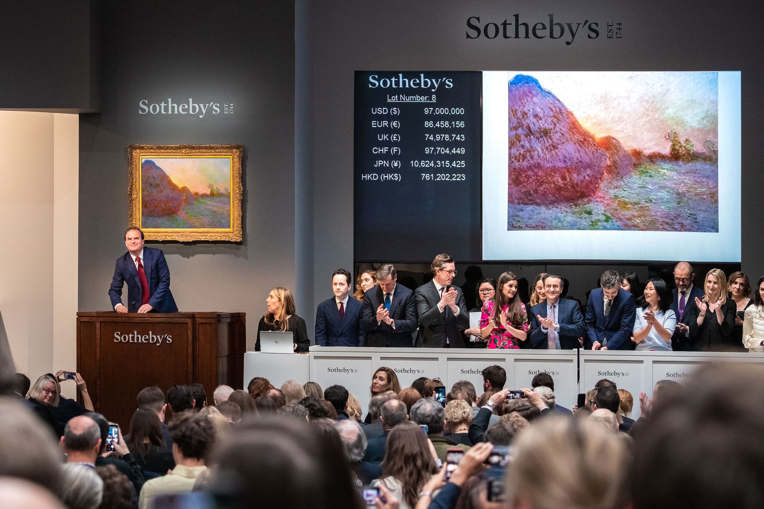 Auction at Sotheby's with auctioneer at podium, painting on display, and audience clapping.