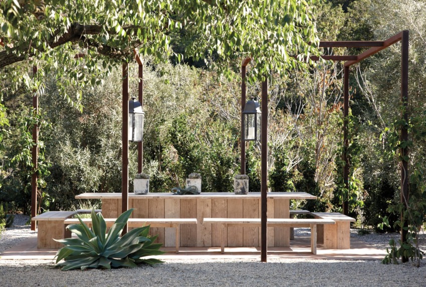 Outdoor wooden dining area with benches and large lanterns under leafy trees, surrounded by greenery and gravel ground.