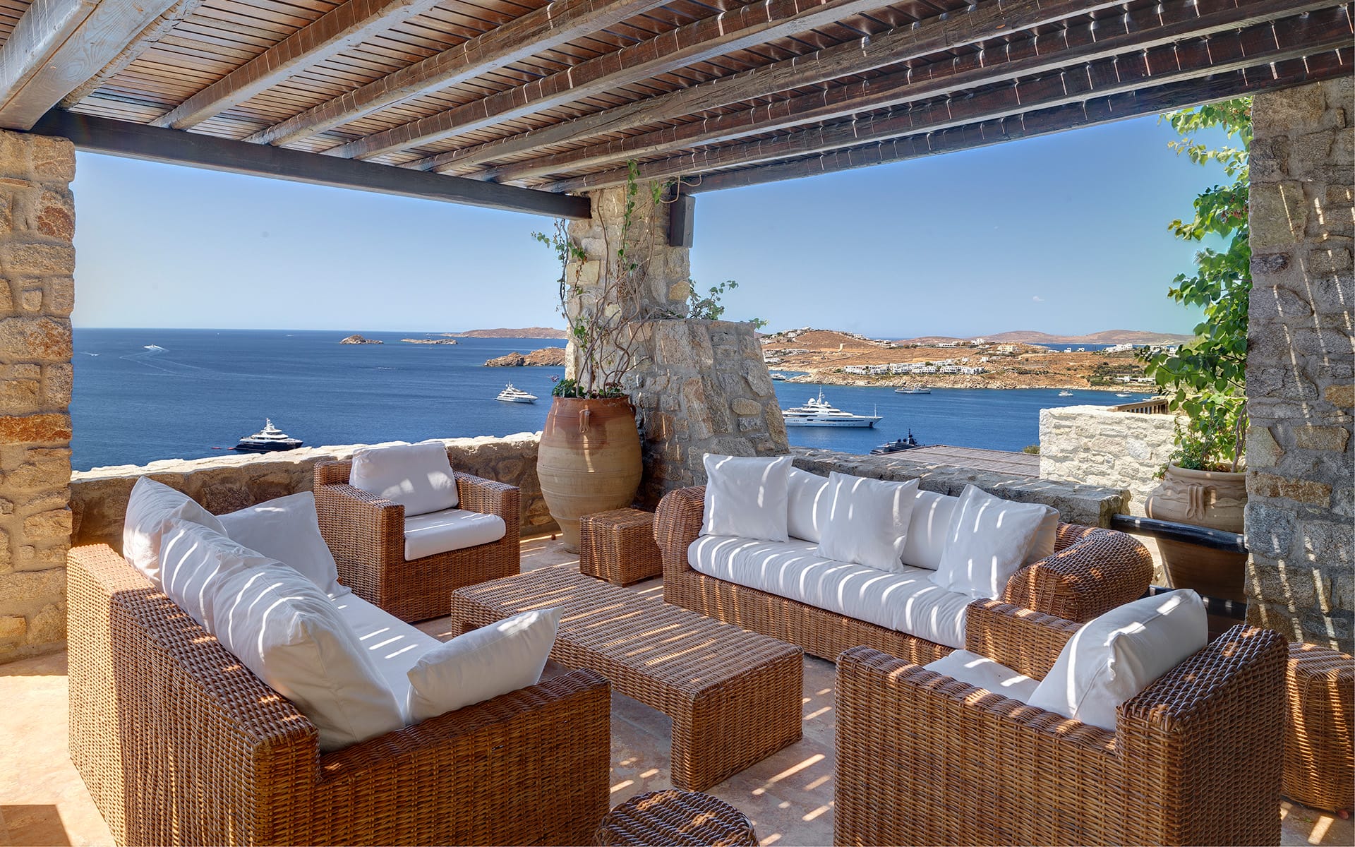 Outdoor patio with wicker furniture overlooking a coastal view with boats and a distant shoreline under a clear blue sky.