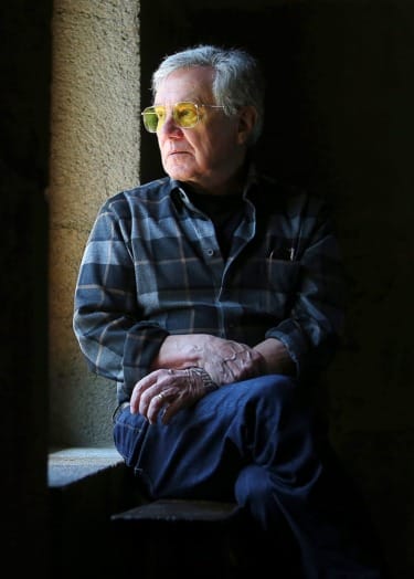 Elderly man wearing yellow glasses and plaid shirt gazing out a window in a dimly lit room, hands resting on his knee.