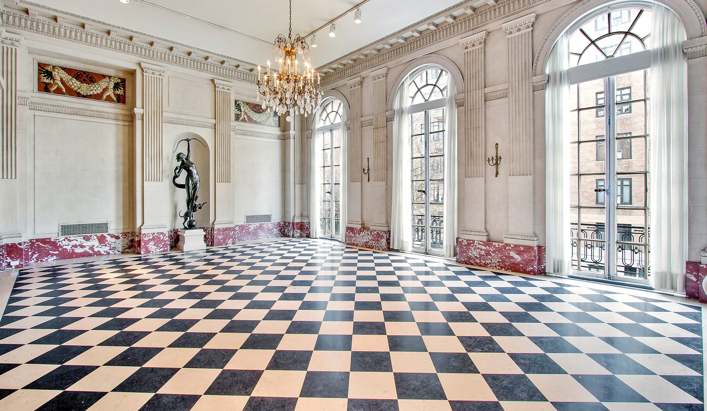 Elegant ballroom with checkered floor, chandelier, large windows, ornate walls, and a statue, filled with natural light.