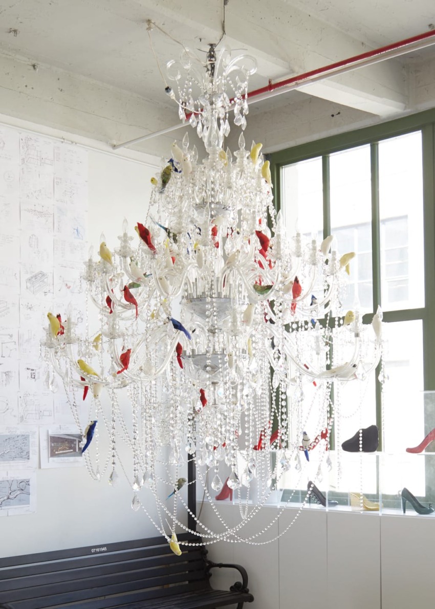 Elegant crystal chandelier adorned with colorful bird ornaments in a bright room with large windows and architectural drawings.