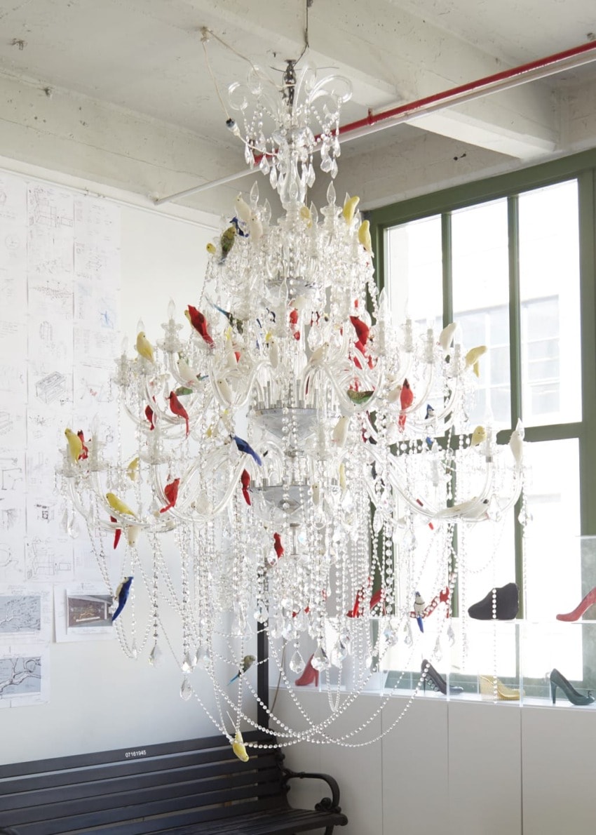 Elegant crystal chandelier adorned with colorful bird ornaments in a bright room with large windows and architectural drawings.