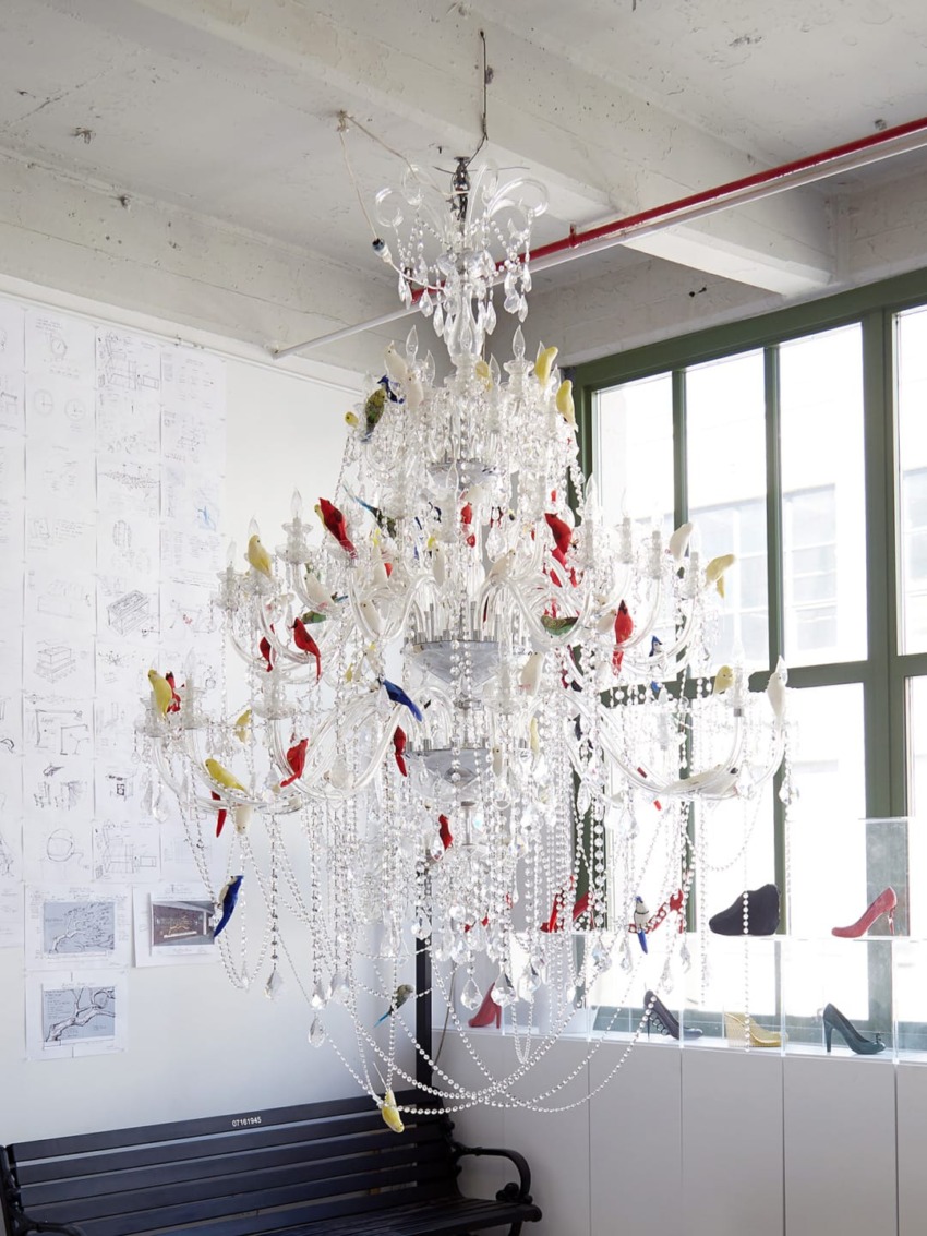 Large ornate chandelier adorned with colorful bird decorations hanging in a modern, industrial-style room with large windows.